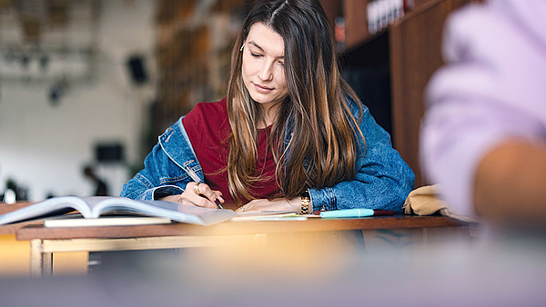 Eine junge Frau in einer Jeansjacke sitzt an einem Tisch und schreibt in ein Notizbuch. Vor ihr liegt ein aufgeschlagenes Buch, und im Hintergrund ist ein unscharfer Raum erkennbar.