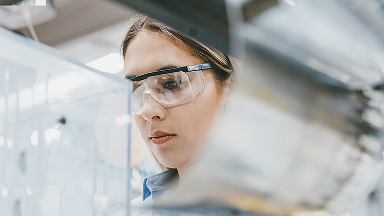  Eine junge Frau mit Schutzbrille arbeitet konzentriert an einer Maschine oder in einem Laborumfeld. Im Hintergrund ist eine industrielle Umgebung erkennbar