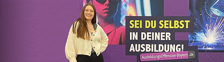 ine junge Frau steht lächelnd vor einer violetten Werbewand mit der Aufschrift „SEI DU SELBST IN DEINER AUSBILDUNG!“ und der Website „AusbildungsOffensive-Bayern.de“. Im Hintergrund sind stilisierte Bilder von Schweißen und urbanen Szenen zu sehen.