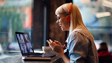 Eine junge Frau mit blonden Haaren und orangefarbenen Kopfhörern sitzt lächelnd vor einem Laptop und nimmt an einem Videoanruf 