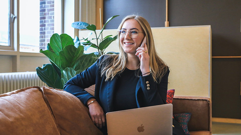 Eine lächelnde Frau in einem dunkelblauen Blazer, die ein Telefonat führt und vor einem offenen silbernen MacBook sitzt. Sie sitzt auf einem beigen Sofa in einem modernen Büro oder Wohnzimmer mit Pflanzen im Hintergrund, was eine freundliche und professionelle Atmosphäre vermittelt.