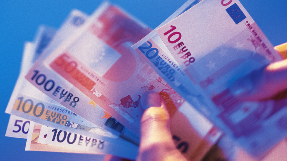 Eine Nahaufnahme von Händen, die mehrere Euro-Geldscheine in verschiedenen Stückelungen, darunter 10, 20, 50 und 100 Euro, halten. Die Szene ist in einem weichen blauen Licht getaucht, das eine professionelle Atmosphäre vermittelt.